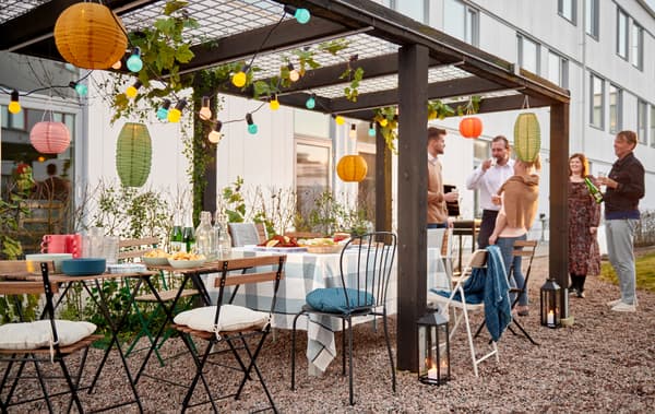 Un gruppo di persone sotto un pergolato decorato a festa accanto a dei tavolini e sedie in un cortile - IKEA