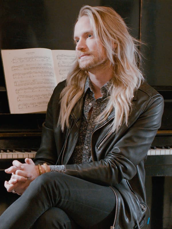 Un hombre con barba rubia y cabello largo rubio, sentado en un taburete junto a un piano.
