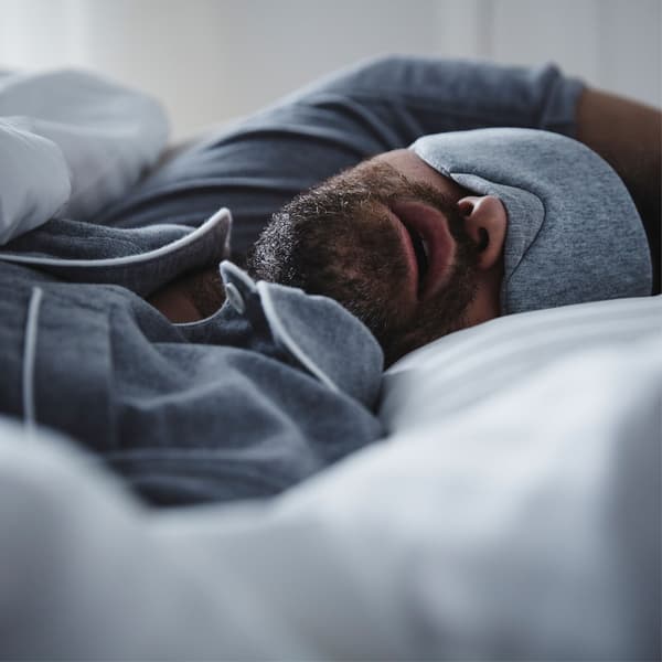 Un hombre duerme profundamente en una cama con un antifaz para no despertarse por la luz exterior.