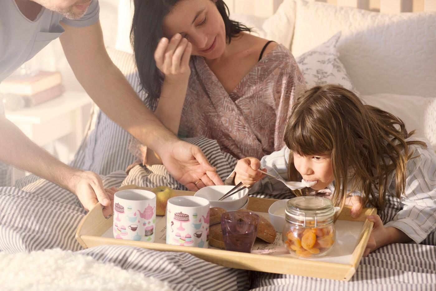 Un hombre, una mujer y una niña tomando desayuno en la cama sobre una bandeja.