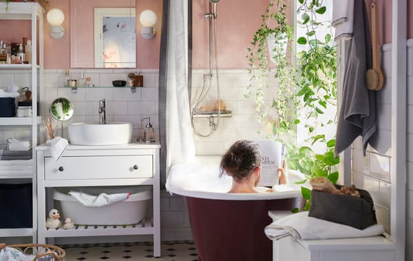 Un home sentado na bañeira nun baño con paredes de cor rosa claro, azulexos brancos, lavabo branco e plantas.