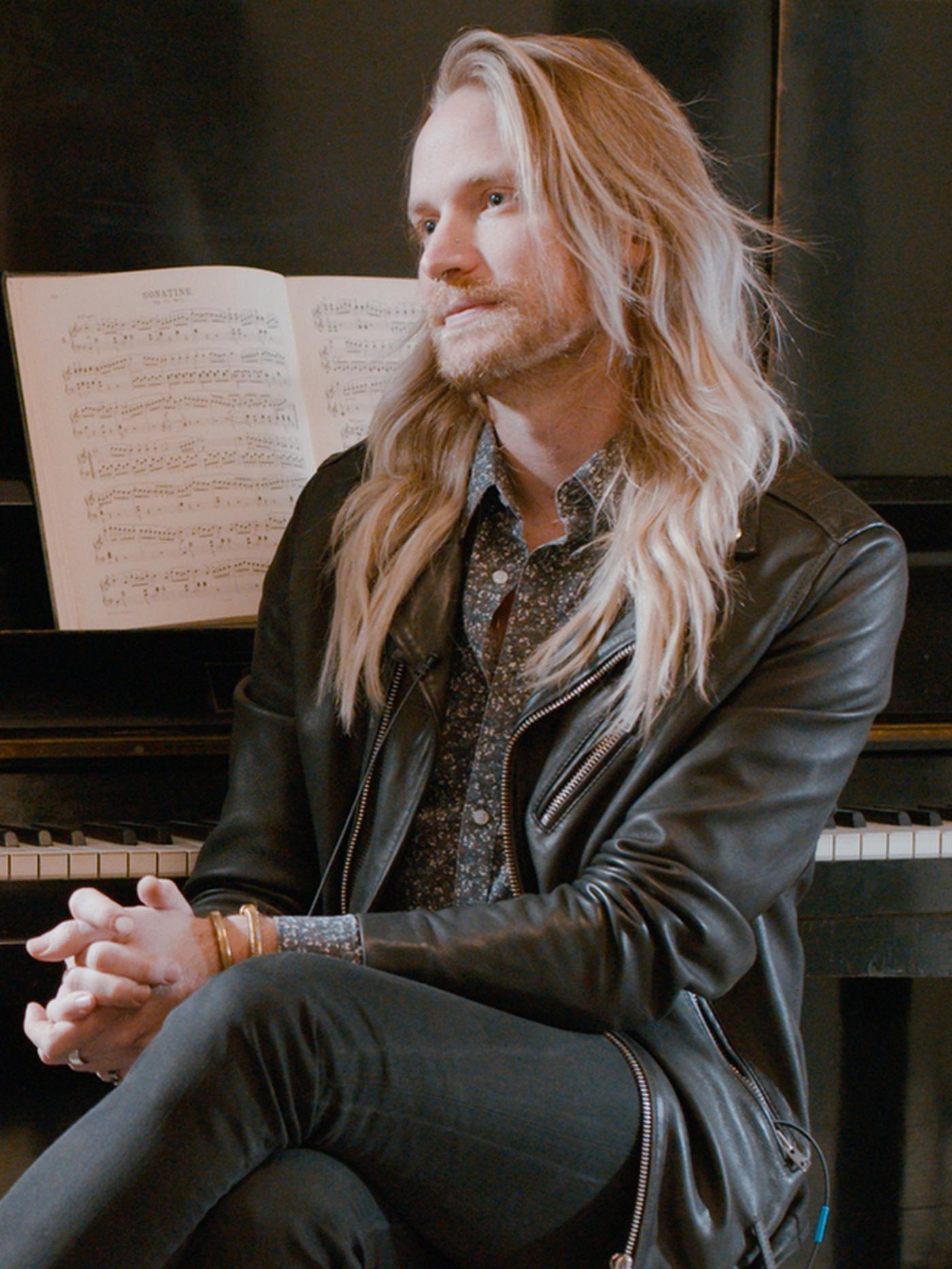 Un homme avec une barbe blonde et de longs cheveux blonds, assis sur un tabouret devant un piano.