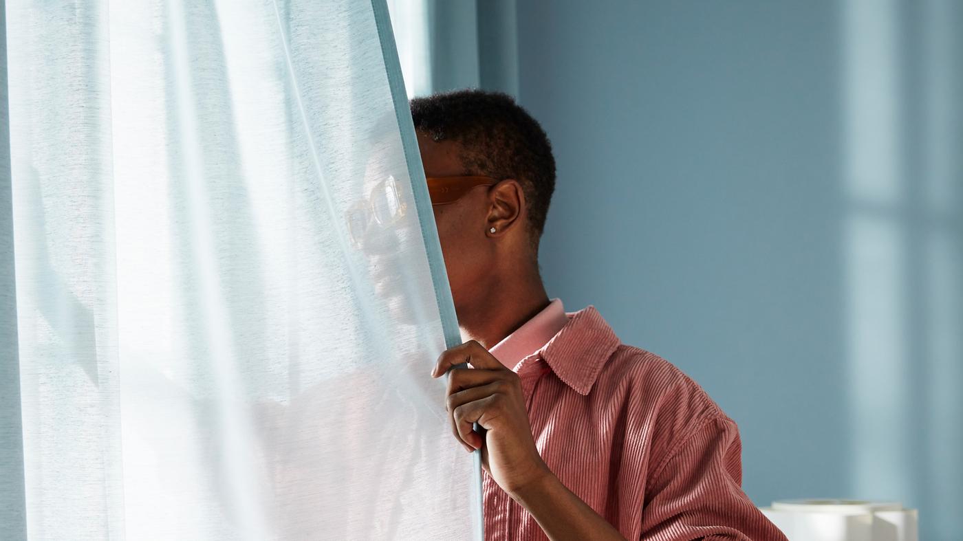 Un homme dans une pièce bleu clair écarte un voilage bleu clair pour regarder par la fenêtre.