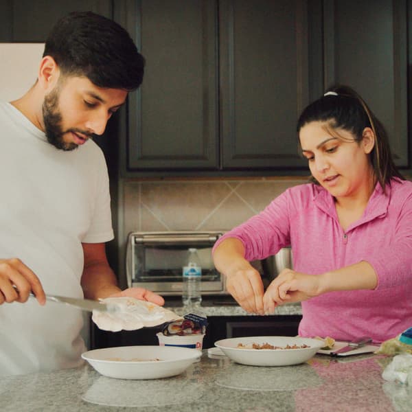Un homme et une femme tenant chacun une assiette préparent le déjeuner dans une cuisine dotée d’un comptoir en pierre et d’armoires grises.