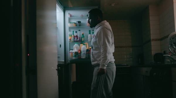Un homme portant un chandail à capuchon blanc et des pantalons de jogging gris se tient debout dans une pièce sombre, et il est en train de regarder ce qui se trouve dans le réfrigérateur.