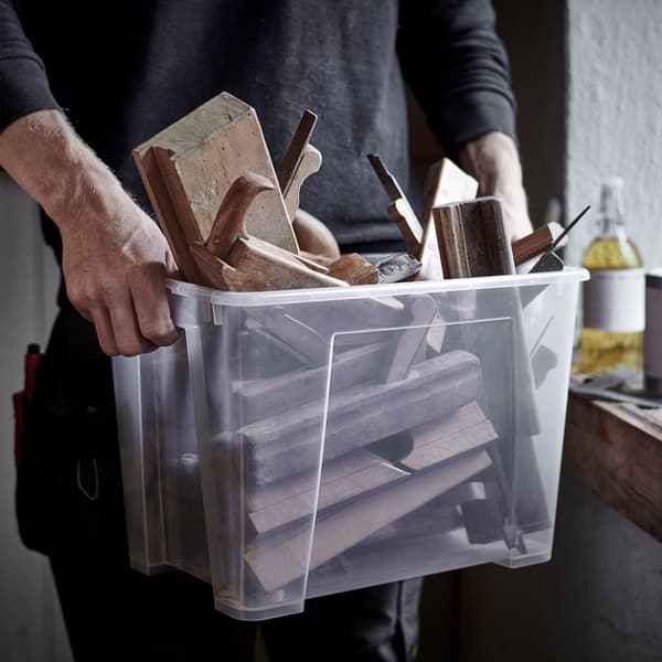 Un homme portant une boîte en plastique transparente SORTERA remplie de morceaux de bois et d’outils en bois.
