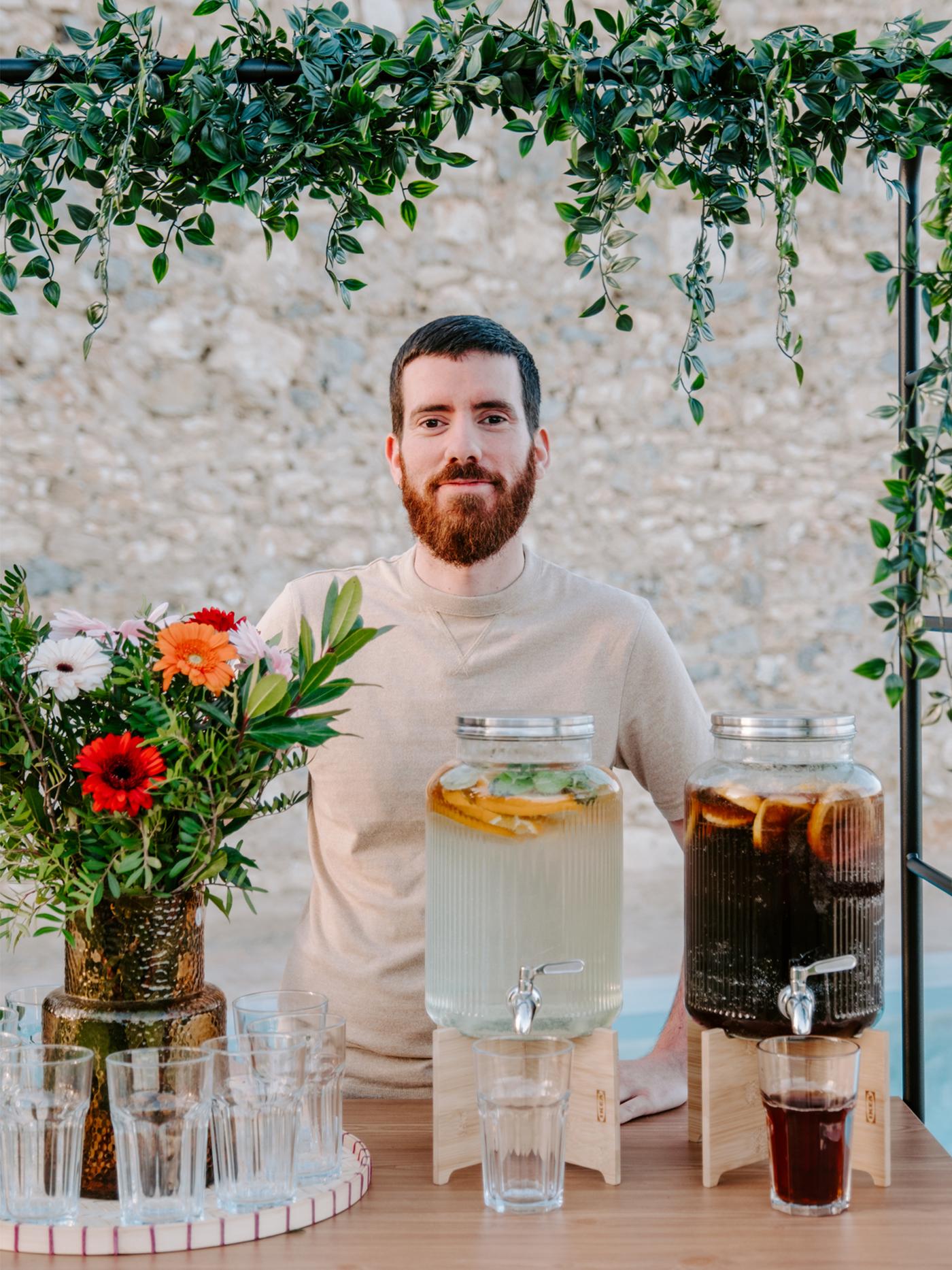 Un homme se tient derrière deux jarres remplies de boissons, avec un vase en verre avec un bouquet de fleurs sur le côté gauche.