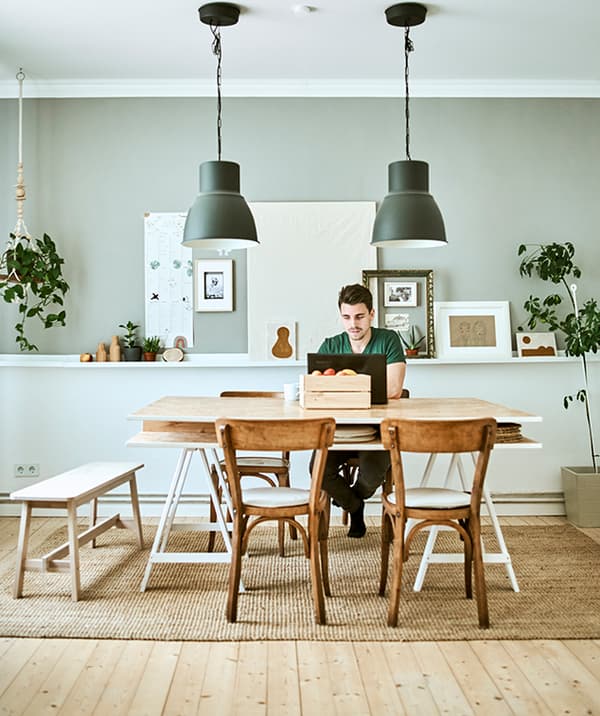 Un homme travaille sur un ordinateur portable sur une table en bois, deux suspensions grises pendent au-dessus et des reproductions sont accrochées au mur gris derrière.
