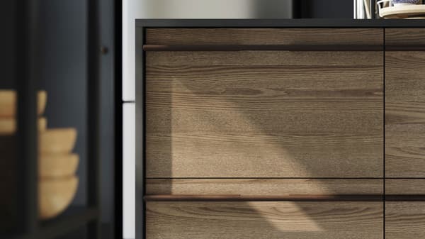 Un îlot de cuisine avec des façades en placage de frêne brun foncé de STENSTA, complété par des tabourets de bar et une grande plante imprimée sur le mur.