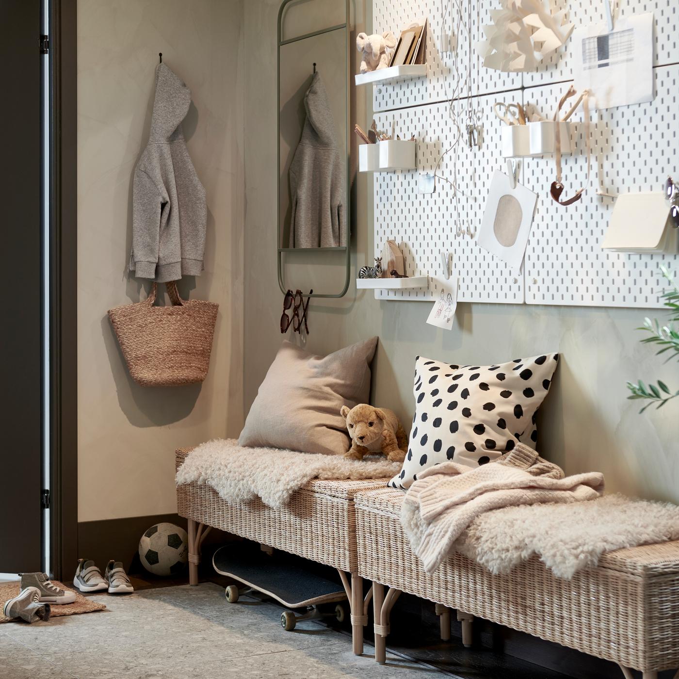 Un ingresso con due panche con contenitore interno in rattan e vari cuscini e tappeti appoggiati sopra, più due pannelli portaoggetti bianchi e uno specchio verde.