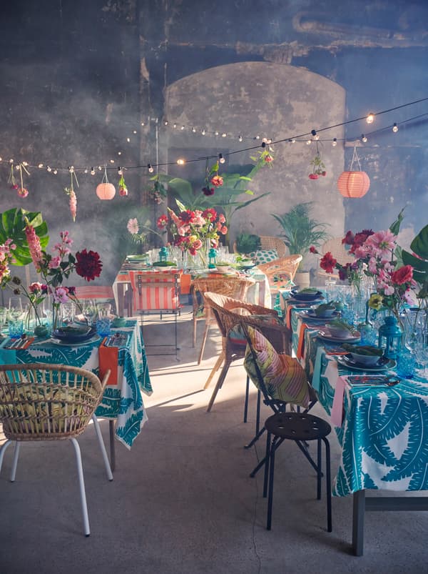 Un intérieur avec des tables dressées et recouvertes de tissus UGGLEMOTT en guise de nappes, surmontées de guirlandes lumineuses SOLVINDEN entrecroisées.