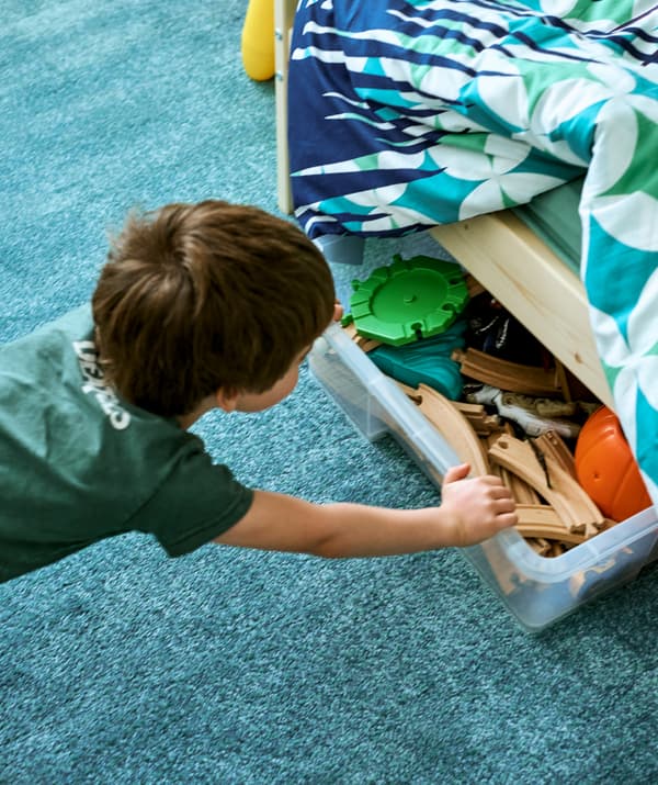 Un jeune garçon pousse une boîte de rangement transparente pleine de rails de petit train sous un lit orné de linge de lit coloré, sur un tapis turquoise.