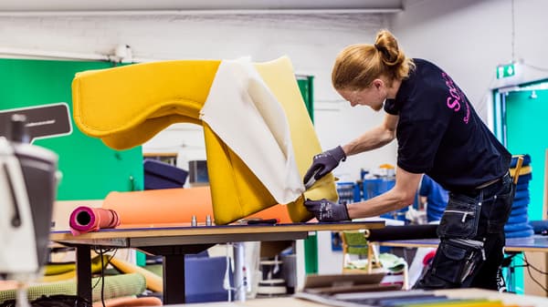 Un jeune homme aux cheveux en queue de cheval rehousse un fauteuil STRANDMON avec du textile jaune.