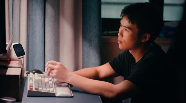 Un jeune homme aux cheveux foncés est assis devant un clavier en train de regarder un écran d'ordinateur dans un éclairage ambiant.