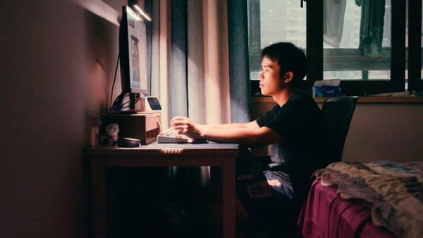 Un jeune homme dans une chambre à coucher sombre d'un apartement urbain est assis près d'un bureau avec un clavier, en train de regarder un écran d'ordinateur.