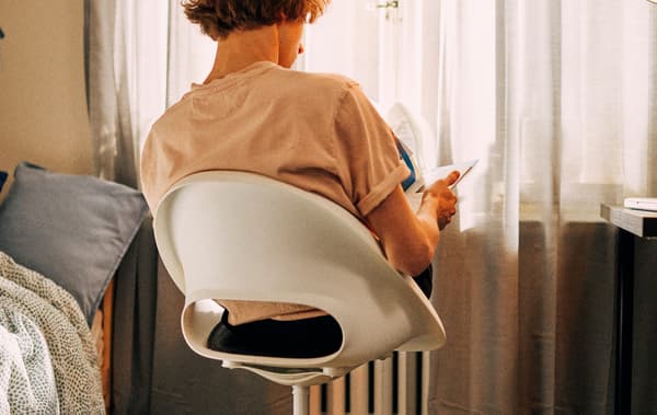 Un jeune homme étudie, assis sur une chaise pivotante devant une fenêtre, les pieds étendus sur un radiateur.
