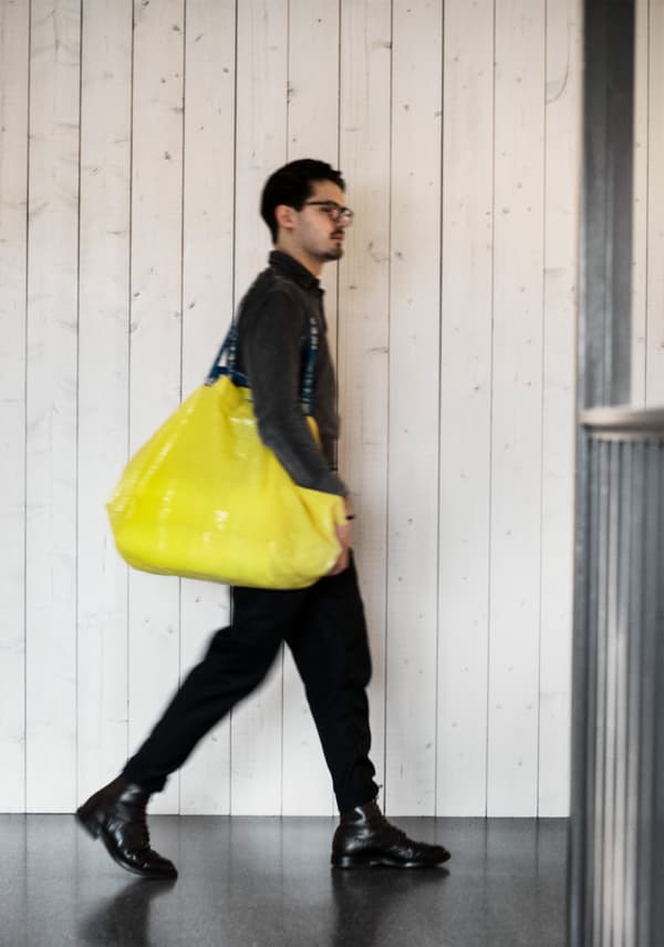 Un jeune homme habillé en noir se baladant dans un magasin IKEA et portant un sac de shopping jaune.