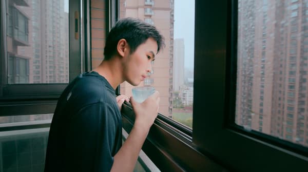 Un jeune homme tient un verre d’eau tout en regardant par la fenêtre d’un appartement.
