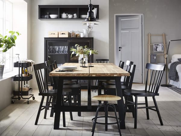Un juego de comedor con una mesa de acacia SKOGSTA y sillas negras NORDVIKEN a su alrededor, un carrito RASKOG cerca y una unidad de almacenamiento con vasos detrás de la mesa