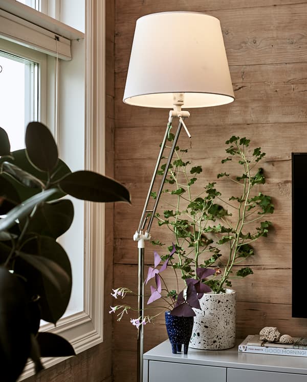 Un lampadaire éclairé avec un abat-jour blanc à côté d’un meuble TV gris avec une plante et des livres au-dessus.