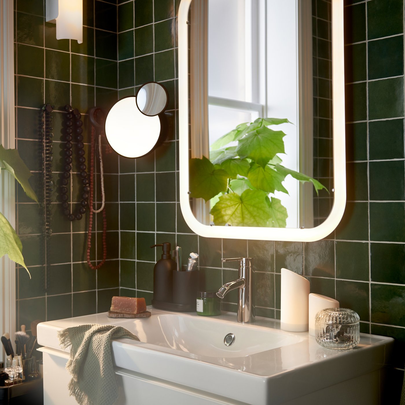 Un lavabo bianco e uno specchio STORJORM con illuminazione integrata in un piccolo bagno con piastrelle verde scuro - IKEA.