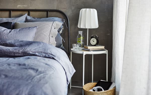 Un letto a fianco di un comodino su cui sono posati un bicchiere d'acqua, libri, una sveglia e una lampada - IKEA