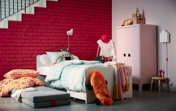 Un letto bianco con biancheria colorata. Sul pavimento, si trova un materssao con piumino e plaid arancioni. Sullo sfondo, c'è una parete di mattoni con una scala con ringhiera in metallo. A lato del letto, un guardaroba rosa con alcuni scatoloni e un appendiabiti.