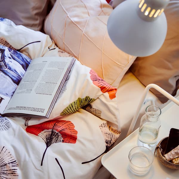 Un letto con biancheria a motivo floreale, cuscini e un libro illuminati da una lampada vicino a un tavolino bianco con una caraffa e un bicchiere d’acqua - IKEA