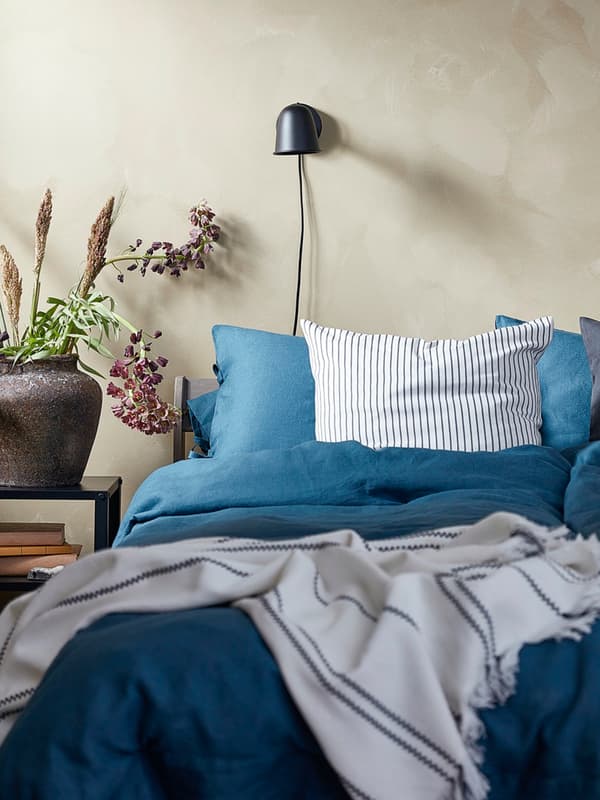 Un letto con biancheria e cuscini sui toni dell'azzuro e del blu. Alla parete, una lampada nera e accanto un tavolino nero con un vaso di fiori.