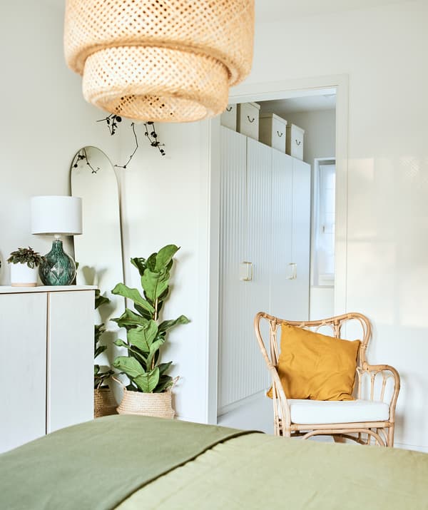 Un letto con biancheria verde in una stanza con una poltrona in rattan, una lampada a sospensione intrecciata e un disimpegno con una fila di armadi bianchi.