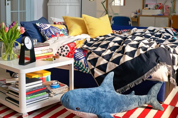 Un letto con cuscini colorati e biancheria a trama geometrica. Sul pavimento c'è un peluche a forma di squalo BLÅHAJ. Accanto al letto, un tavolino bianco TINGBY con libri impilati, un vaso di fiori e una sveglia.