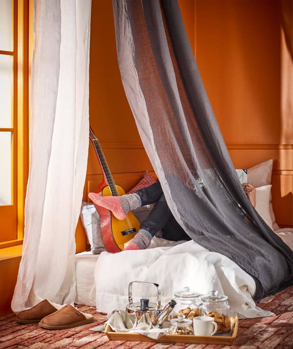 Un letto con materasso e circondato da  tende LEJONGAP fino al pavimento. Sul letto è stesa una persona con accanto una chitarra e dei cuscini. Sul pavimento è posato un vassoio con una teiera, contenitori di vetro dentro dei biscotti e una tazza.