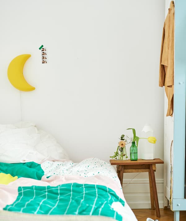 Un letto con piumino verde, rosa e giallo contro una parete con una lampada gialla a forma di luna; accanto, un tavolino in legno con sopra dei vasi.