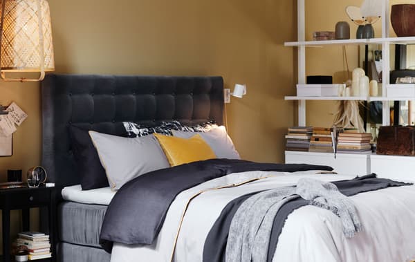 Un letto con una testiera trapuntata di velluto grigio e diversi strati di tessili. Accanto una libreria e un comodino nero - IKEA