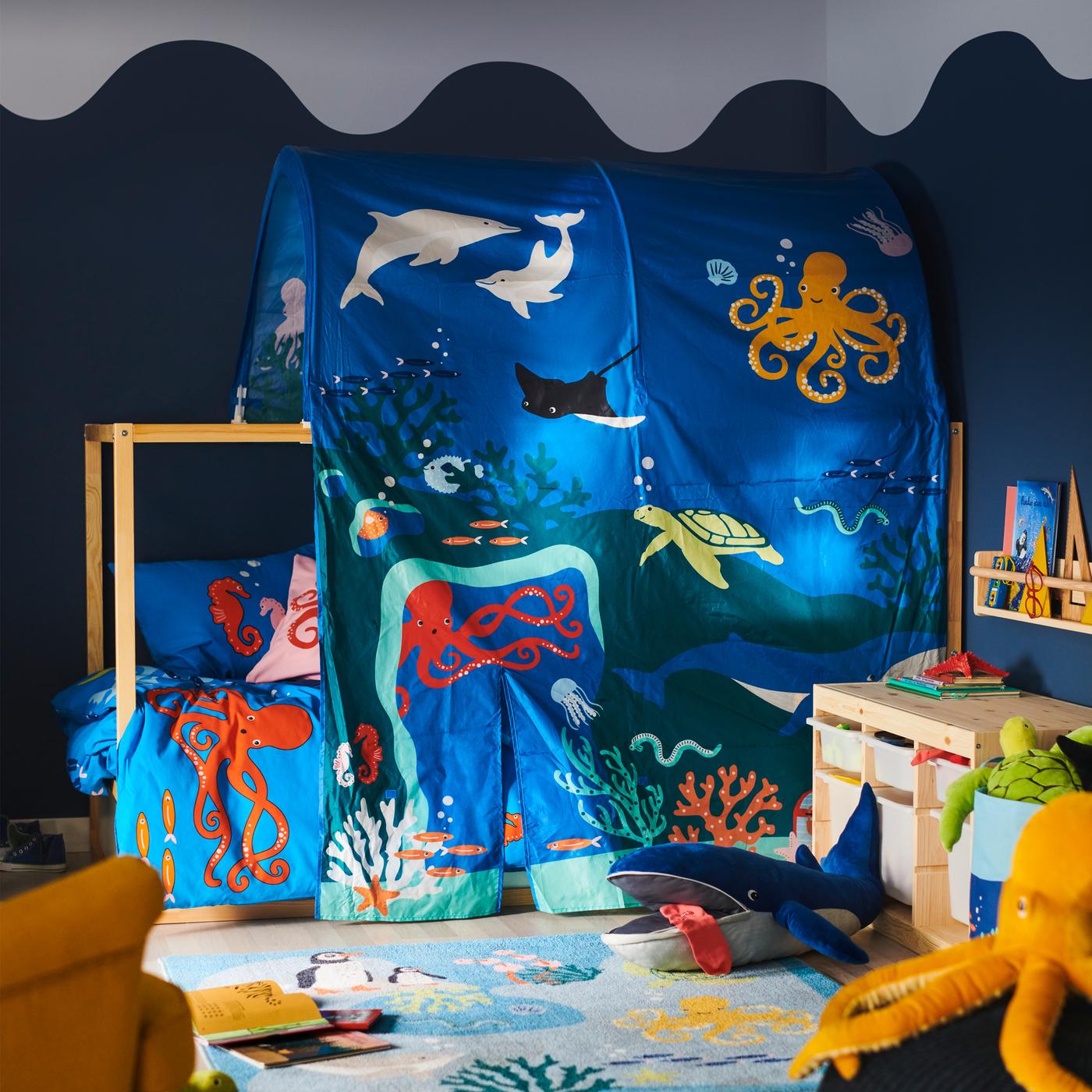 Un letto coperto con una tenda per letto KURA con motivo di animali dell'oceano; di fronte, un giocattolo morbido BLÅVINGAD a forma di balenottera azzurra su un tappeto BLÅVINGAD - IKEA.