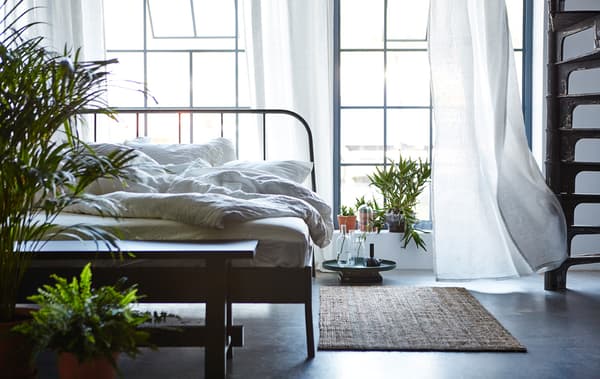 Un letto disfatto in una camera con le finestre aperte, un tappeto e molte piante verdi - IKEA
