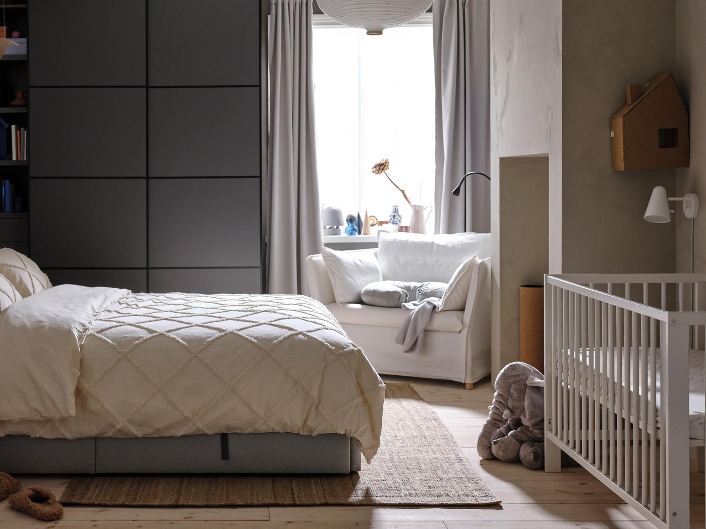 Un letto imbottito GLADSTAD con biancheria da letto NYPONLUGGMAL, di fronte a un guardaroba PAX/MEHAMN grigio scuro e a una poltrona bianca.