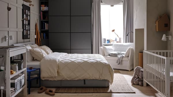 Un letto imbottito grigio, con biancheria NYPONLUGGMAL, di fronte a un guardaroba PAX/MEHAMN grigio scuro e a una poltrona bianca.