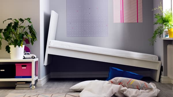 Un letto inserito in una nicchia. Il letto risulta troppo grande. Sul pavimento, cuscino e biancheria da letto. A lato, un tavolino von libri, giochi, contenitori e una pianta in vaso.