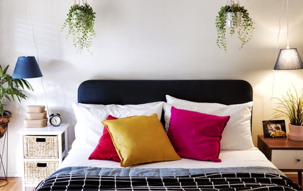 Un letto matrimoniale con cuscini colorati e strati di coperte, due comodini, lampade a sospensione e vasi appesi.
