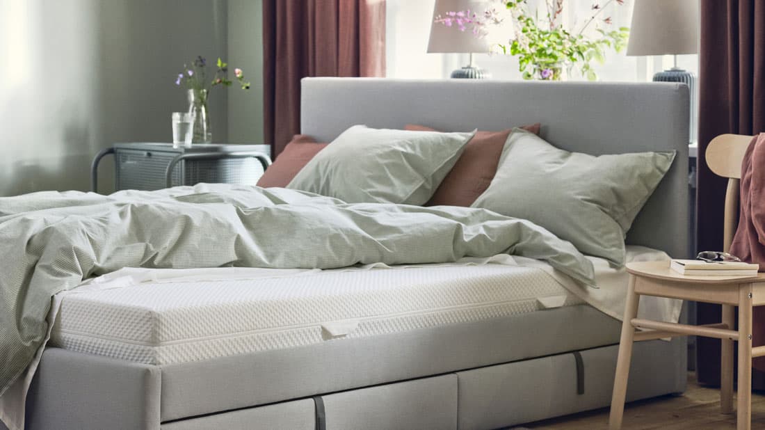 Un letto matrimoniale imbottito grigio con accanto una sedia utilizzata come comodino. Dietro alla testata una finestra con delle tende rosa antico.