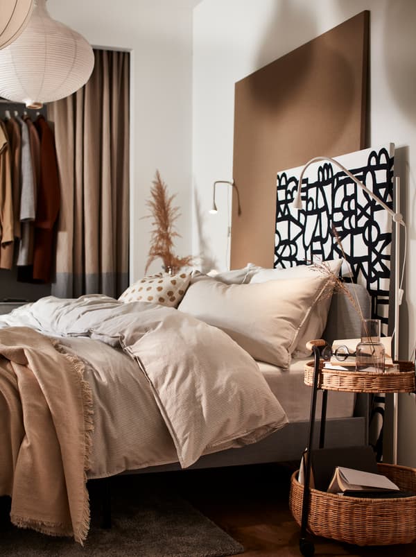 Un lit avec du linge de lit beige et un panneau mural réalisé à partir d’un panneau de particules couvert de tissu au mètre SKUGGBRÄCKA noir et blanc.