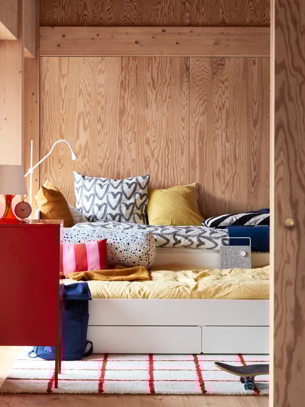 Un lit avec lit tiroir SLÄKT dans une chambre à coucher aux murs revêtus de panneaux de bois, habillé d’une couette avec housse jaune et d’une autre avec housse blanche à motifs de cœurs gris.