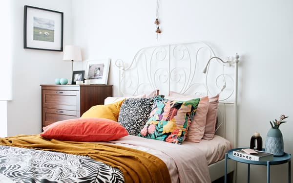Un lit avec tête de lit blanche en fer forgé, une couette rose, un plaid jaune foncé et des coussins en rose, orange, doré et monochrome.