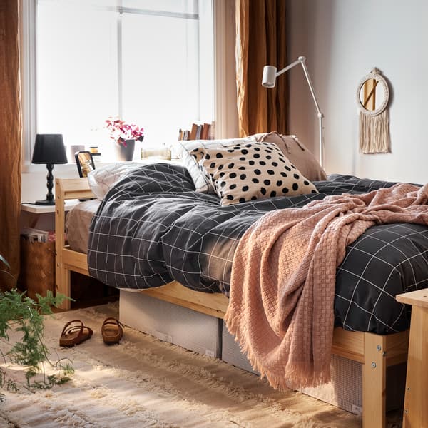 Un lit avec un cadre en pin et du linge de lit à carreaux noirs, plus des caisses de rangement blanches/grises et un tapis en dessous.