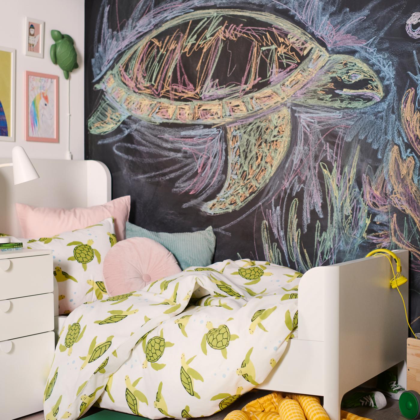 Un lit BUSUNGE est habillé d’une housse de couette BLÅVINGAD à motifs de tortue verte avec des coussins devant un mur tableau noir.