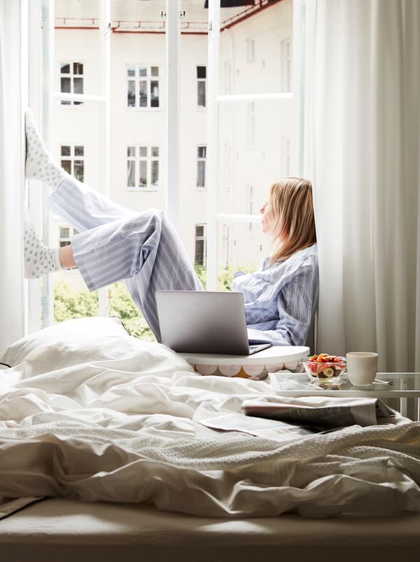 Un lit défait à côté d’une fenêtre ouverte au soleil. Une femme est assise sur l’appui de fenêtre avec, à côté d’elle, un ordinateur portable posé sur un support BYLLAN.