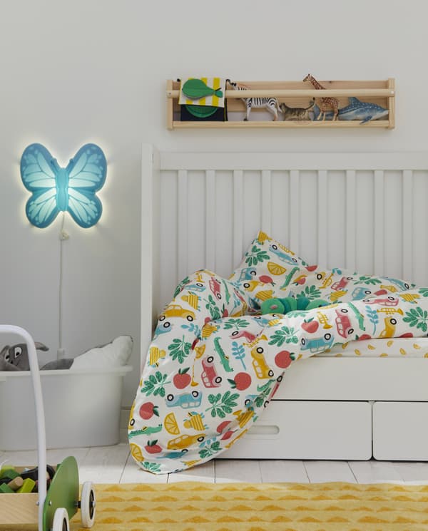 Un lit d’enfant blanc avec linge de lit à motif multicolore, une applique en forme de papillon, un tapis jaune et des jouets rangés sur une étagère.