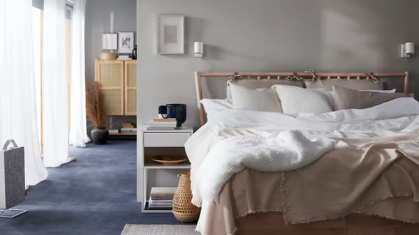 Un lit en bouleau BJÖRKSNÄS avec un linge de lit blanc et beige à côté d’une table de chevet blanche, d’une lampe de table en bambou et d’un purificateur d’air.