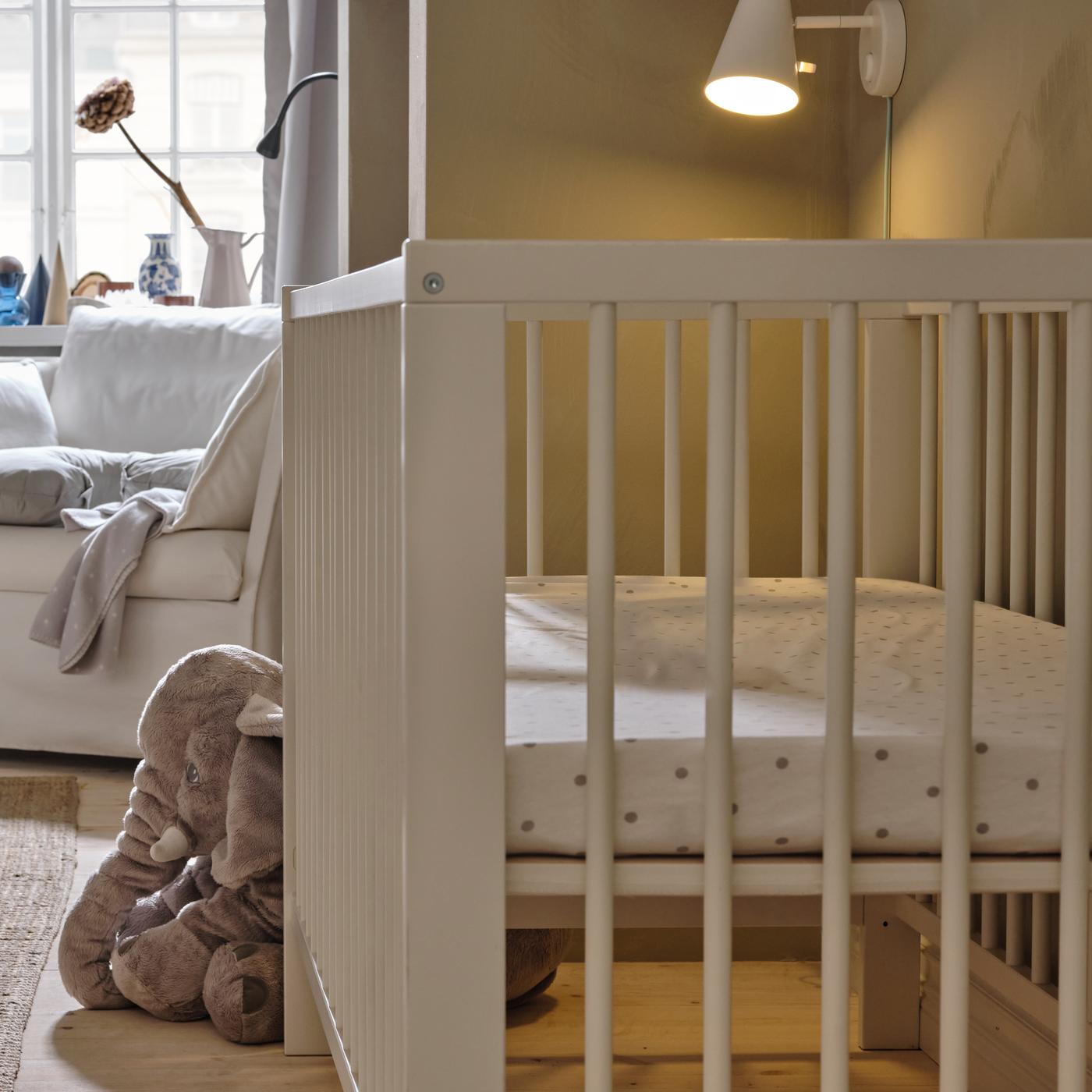 Un lit enfant GULLIVER blanc sur lequel est posé un éléphant en peluche. Il y a un fauteuil blanc à l’arrière. Une applique est allumée à côté.